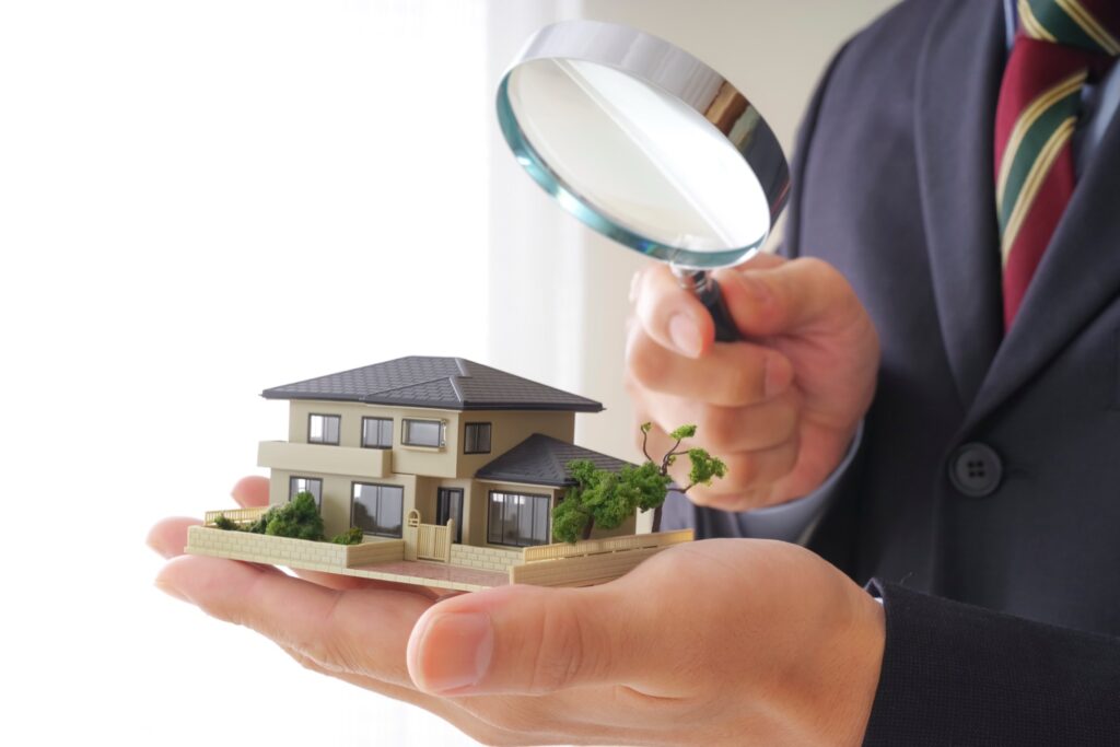 手のひらに乗った住宅模型を虫眼鏡で細部まで確認するスーツ姿の人物。施工事例における住宅の品質検査や詳細評価の様子。