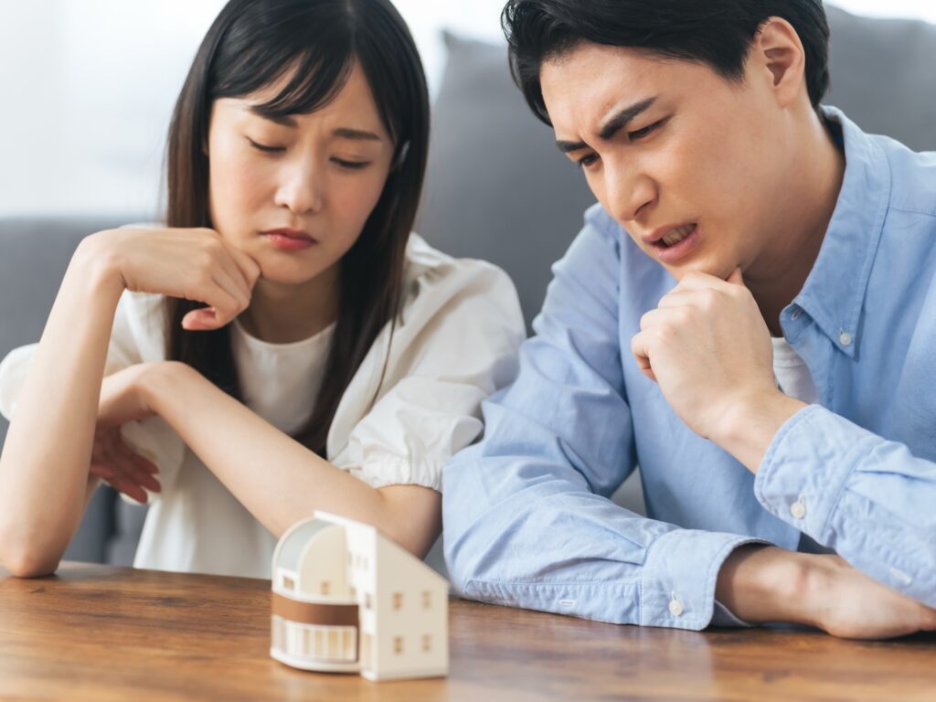 深刻な表情で住宅模型を見つめ、家の売却や相続について考え込む若い夫婦。空き家を放置することのリスクなど真剣に悩んでいる様子。