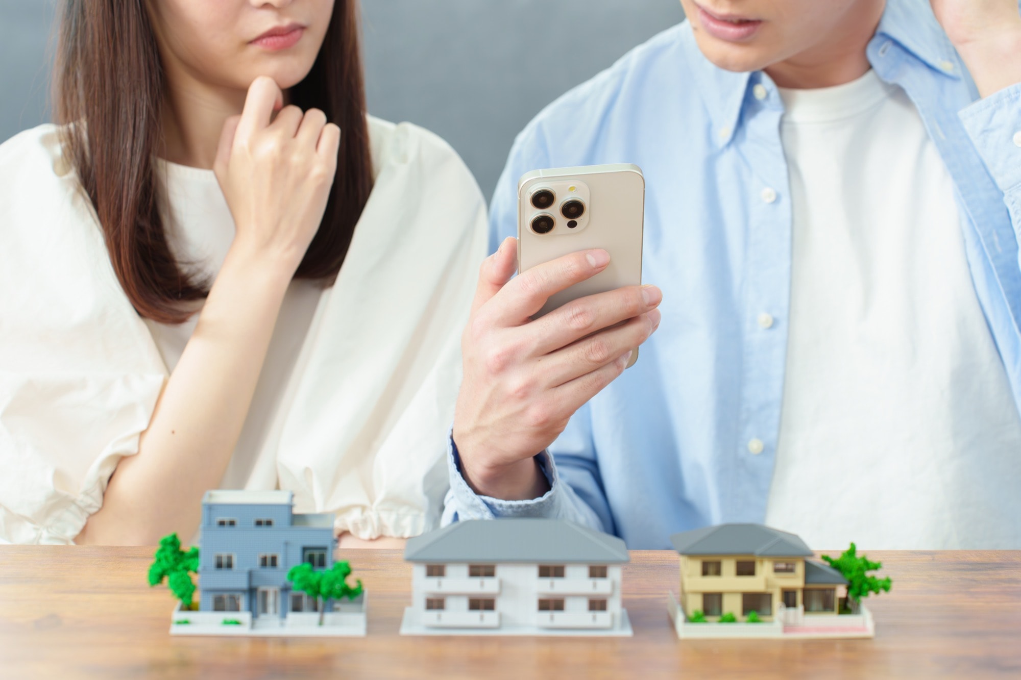 住宅の売却を検討する夫婦が、住宅模型を前にスマートフォンで情報収集し相談する様子