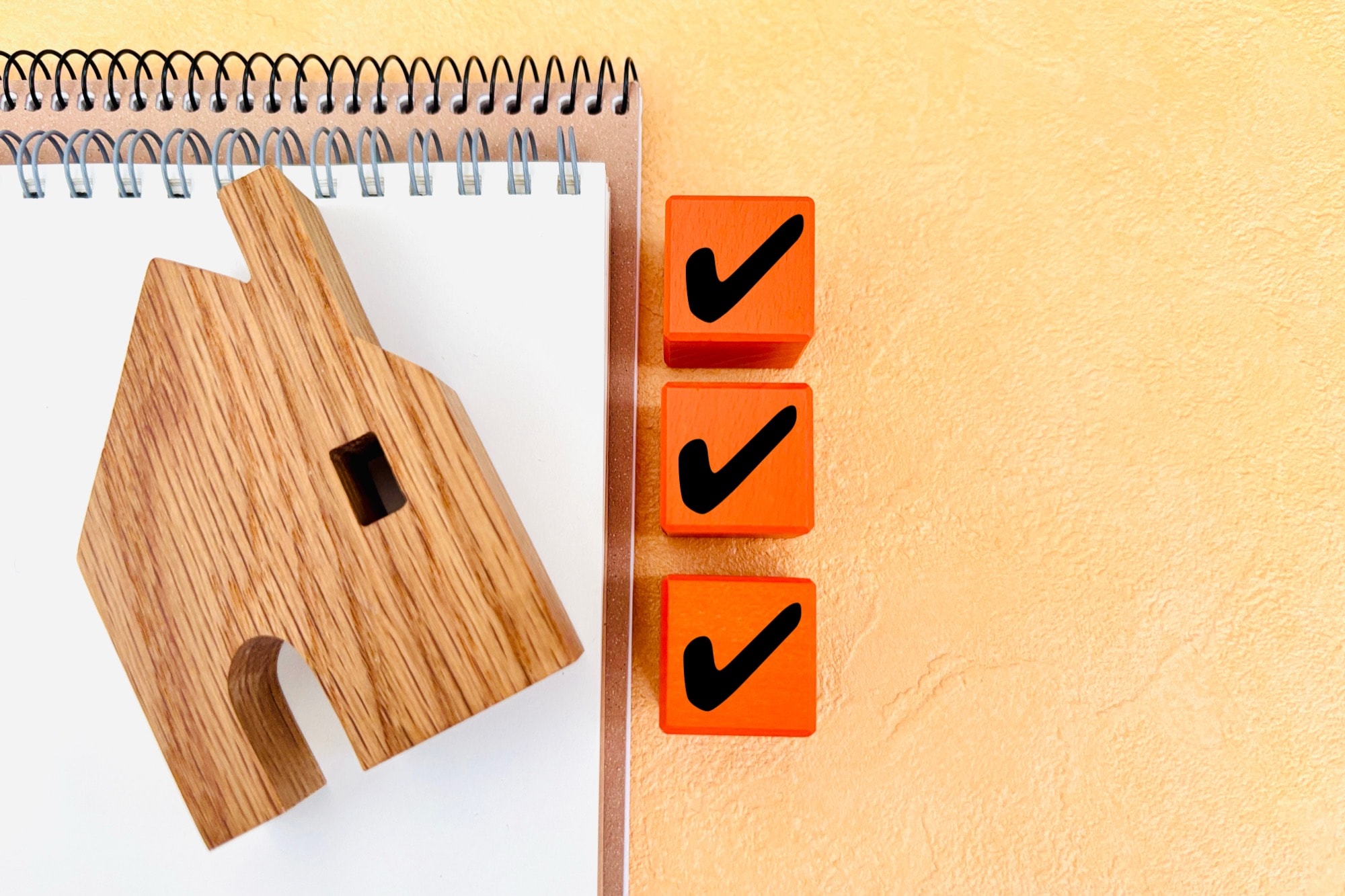 木製の家の模型とノート、チェックマークが書かれたオレンジ色のブロック。住宅売却の計画、不動産タスクの完了などを象徴。