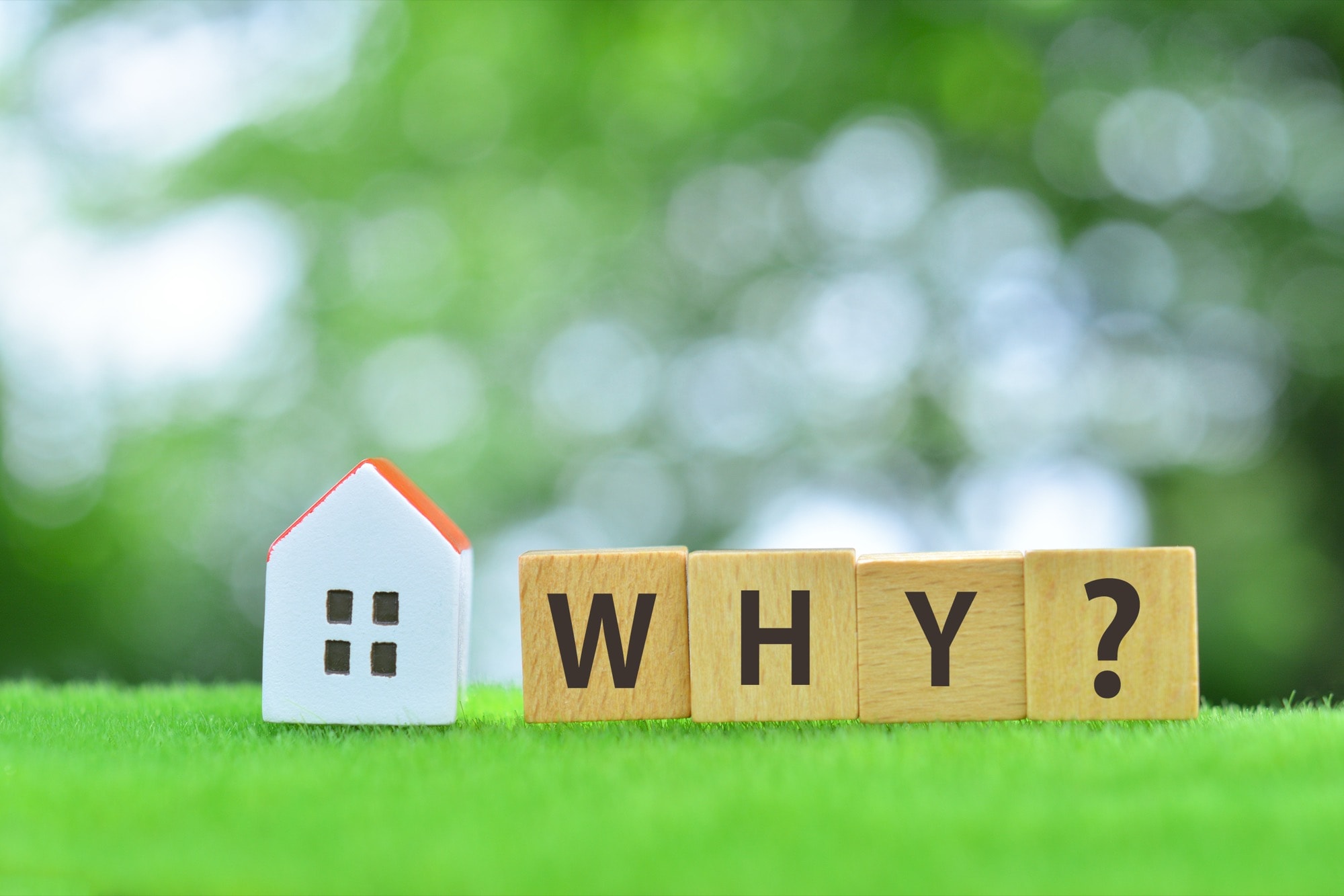 鮮やかな緑の芝生に、白い壁とオレンジの屋根のミニチュアハウスと「WHY?」の文字が書かれた木製ブロック。住宅売却、不動産に対する疑問を表現。