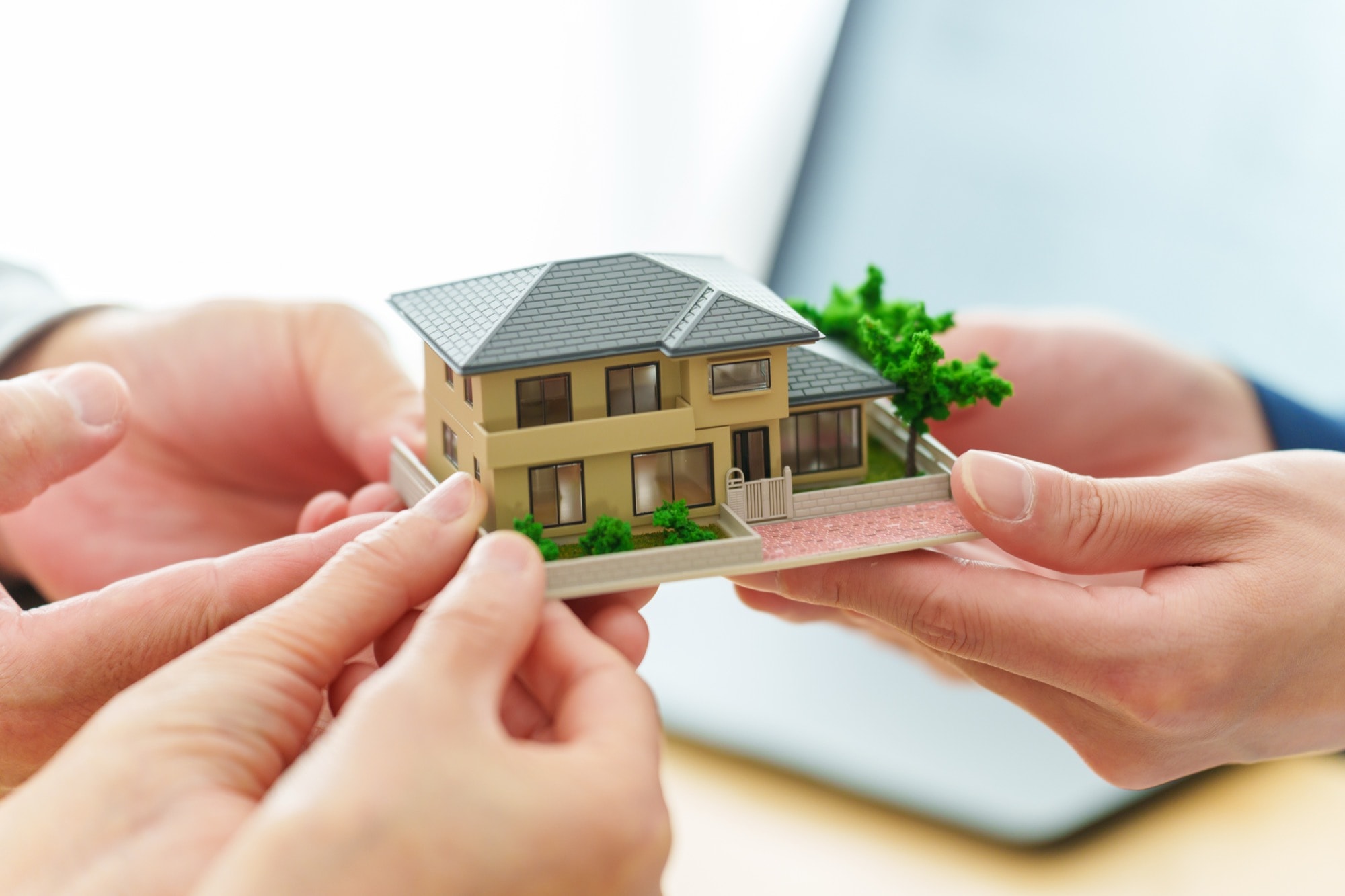 複数の人の手が、庭付きの一軒家を模した精巧なミニチュア模型を大切に支えている。住宅購入、住宅売却、不動産契約、住宅引き渡しにおける安心感とサポートを象徴。
