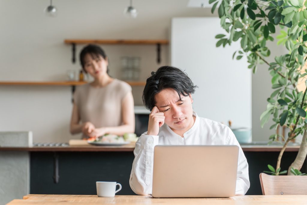 家でパソコンと向き合いながら悩む男性と料理する女性